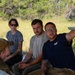 Four individuals are gathered outdoors in what appears to be a briefing or instructional setting near a metal table or bench. One person wearing a navy blue shirt and glasses is gesturing and appears to be explaining or pointing something out to the group. The setting includes tall grass and trees in the background, suggesting a rural or training area.