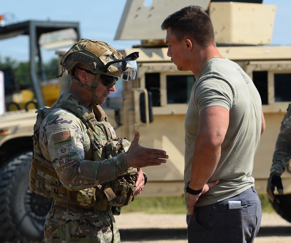 Two military personnel are engaged in a conversation at what appears to be an outdoor military installation or training area. The individual on the left is in full combat gear including OCP (Operational Camouflage Pattern) uniform, tactical helmet, body armor, and sunglasses, while the person on the right is in a sage green t-shirt and dark pants. A tan military HMMWV (Humvee) is visible in the background.