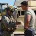 Two military personnel are engaged in a conversation at what appears to be an outdoor military installation or training area. The individual on the left is in full combat gear including OCP (Operational Camouflage Pattern) uniform, tactical helmet, body armor, and sunglasses, while the person on the right is in a sage green t-shirt and dark pants. A tan military HMMWV (Humvee) is visible in the background.