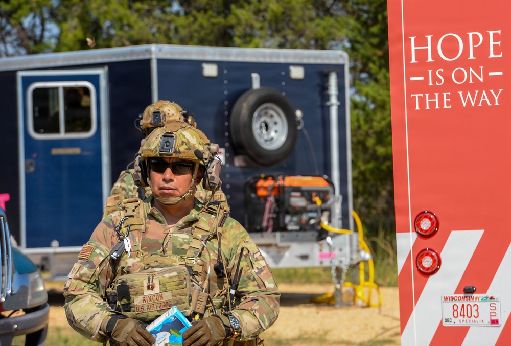 A U.S. Air Force service member named Rincon, wearing full combat gear including a tactical helmet with communications equipment and OCP camouflage uniform, stands in front of a utility trailer and equipment. To the right is a red vehicle or trailer bearing a Wisconsin license plate (8403, Special) and the text 'HOPE IS ON THE WAY,' suggesting a disaster relief or humanitarian assistance operation. The setting appears to be an outdoor staging area with trees in the background.