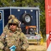 A U.S. Air Force service member named Rincon, wearing full combat gear including a tactical helmet with communications equipment and OCP camouflage uniform, stands in front of a utility trailer and equipment. To the right is a red vehicle or trailer bearing a Wisconsin license plate (8403, Special) and the text 'HOPE IS ON THE WAY,' suggesting a disaster relief or humanitarian assistance operation. The setting appears to be an outdoor staging area with trees in the background.