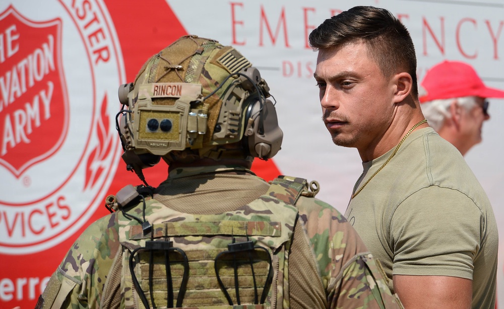 Two military personnel are engaged in conversation in front of a Salvation Army Emergency Disaster Services vehicle. One individual is fully equipped with tactical gear, a combat helmet with communications equipment, and a name tape reading 'RINCON,' while the other wears a military t-shirt and a gold chain. A third person wearing a red cap is partially visible in the background.