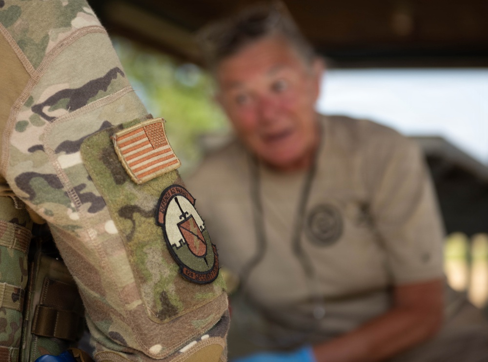 A close-up photograph focusing on the sleeve of a U.S. military uniform in OCP (Operational Camouflage Pattern), displaying an American flag patch and a unit insignia patch. In the blurred background, an older civilian male is visible, appearing to be engaged in conversation. The depth of field technique draws attention to the uniform patches as the primary subject.