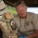 A close-up photograph focusing on the sleeve of a U.S. military uniform in OCP (Operational Camouflage Pattern), displaying an American flag patch and a unit insignia patch. In the blurred background, an older civilian male is visible, appearing to be engaged in conversation. The depth of field technique draws attention to the uniform patches as the primary subject.