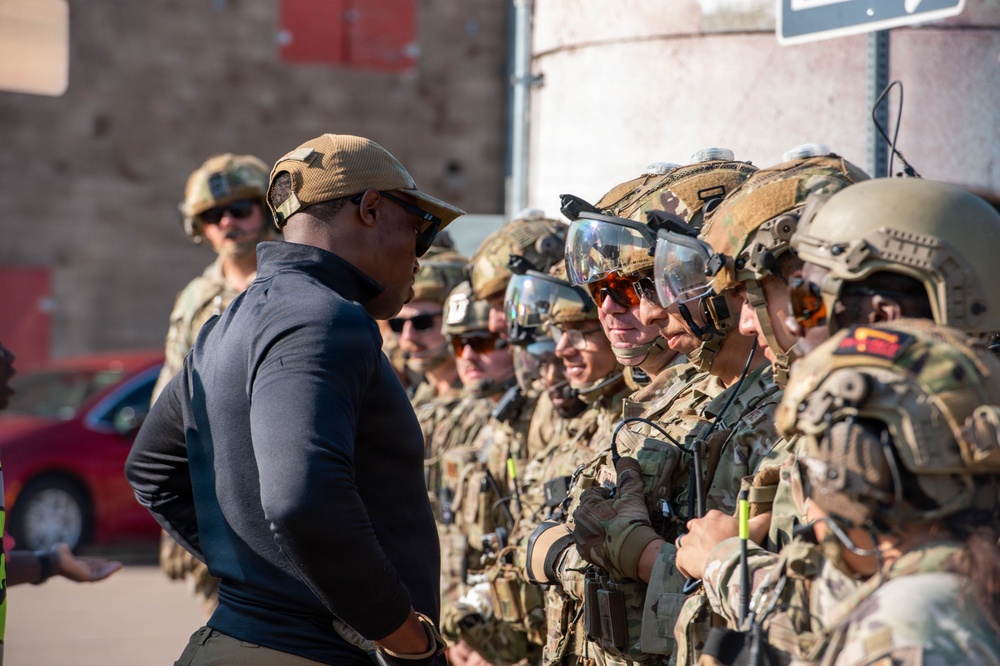 A person in civilian-style tactical clothing and a tan cap appears to be briefing or inspecting a line of fully equipped military or special operations personnel in multicam uniforms. The soldiers are wearing ballistic helmets with mounted visors, tactical vests, and communications equipment, suggesting a pre-mission briefing or inspection. The setting appears to be an outdoor urban training or operational environment.