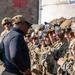 A person in civilian-style tactical clothing and a tan cap appears to be briefing or inspecting a line of fully equipped military or special operations personnel in multicam uniforms. The soldiers are wearing ballistic helmets with mounted visors, tactical vests, and communications equipment, suggesting a pre-mission briefing or inspection. The setting appears to be an outdoor urban training or operational environment.