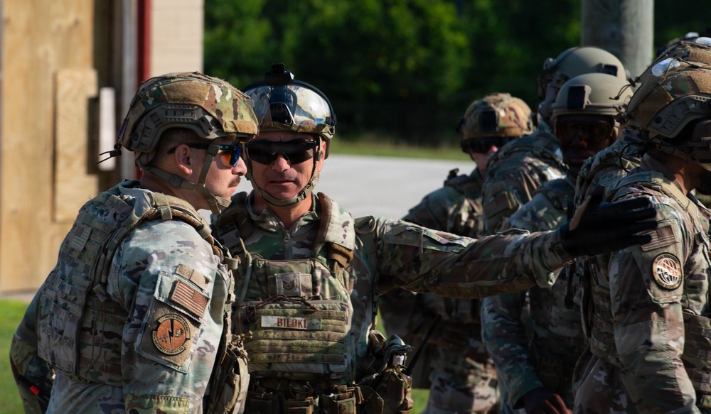 A group of military personnel in full tactical gear are gathered outdoors, appearing to conduct a briefing or coordination discussion. The soldiers are wearing OCP (Operational Camouflage Pattern) uniforms with plate carriers, tactical helmets, and ballistic eyewear. One individual's name tape reads 'BILOKI' and unit patches are visible on their shoulders.