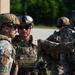 A group of military personnel in full tactical gear are gathered outdoors, appearing to conduct a briefing or coordination discussion. The soldiers are wearing OCP (Operational Camouflage Pattern) uniforms with plate carriers, tactical helmets, and ballistic eyewear. One individual's name tape reads 'BILOKI' and unit patches are visible on their shoulders.