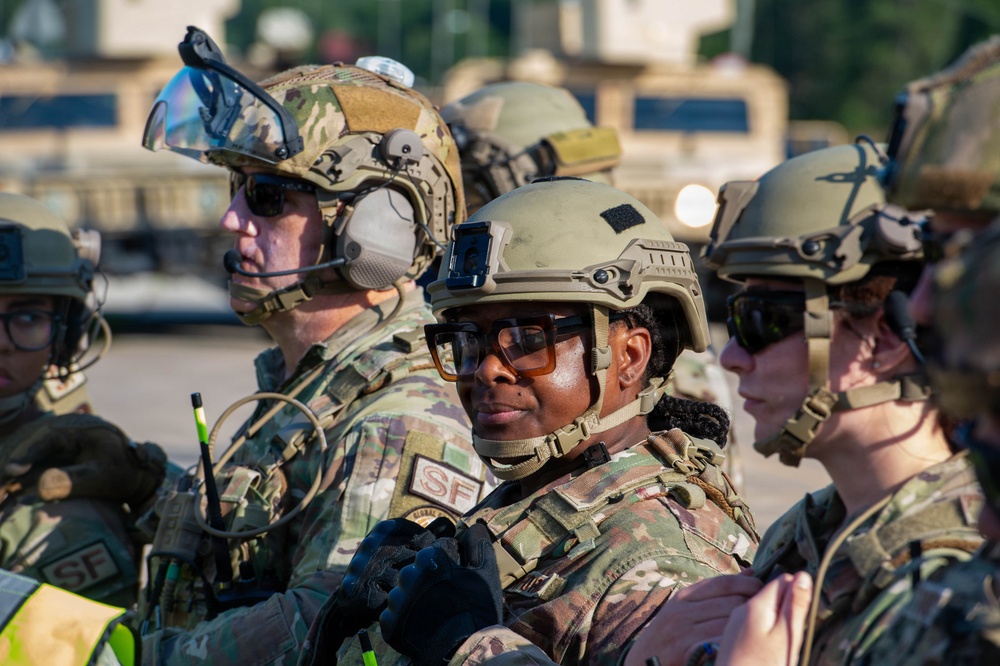 A group of military personnel in full tactical gear are assembled together, likely during a training exercise or operational briefing. Multiple soldiers wearing OCP (Operational Camouflage Pattern) uniforms are visible, with 'SF' patches indicating Security Forces. The central figure faces the camera while others are oriented in various directions.