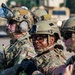 A group of military personnel in full tactical gear are assembled together, likely during a training exercise or operational briefing. Multiple soldiers wearing OCP (Operational Camouflage Pattern) uniforms are visible, with 'SF' patches indicating Security Forces. The central figure faces the camera while others are oriented in various directions.
