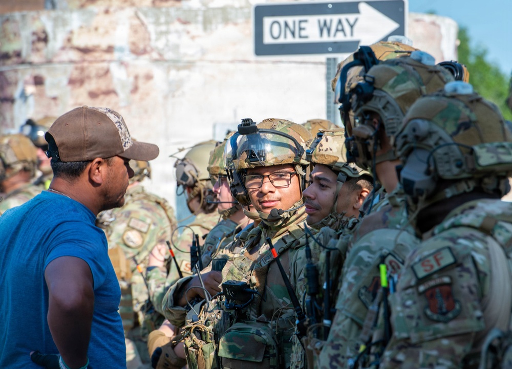 A group of fully equipped military personnel in camouflage uniforms and tactical gear are engaged in conversation with a civilian man wearing a blue shirt and brown cap. The soldiers are wearing combat helmets, tactical vests, communications equipment, and patches including 'SF' (Security Forces). The scene appears to take place in an urban training or operational environment near a 'ONE WAY' street sign.