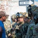 A group of fully equipped military personnel in camouflage uniforms and tactical gear are engaged in conversation with a civilian man wearing a blue shirt and brown cap. The soldiers are wearing combat helmets, tactical vests, communications equipment, and patches including 'SF' (Security Forces). The scene appears to take place in an urban training or operational environment near a 'ONE WAY' street sign.