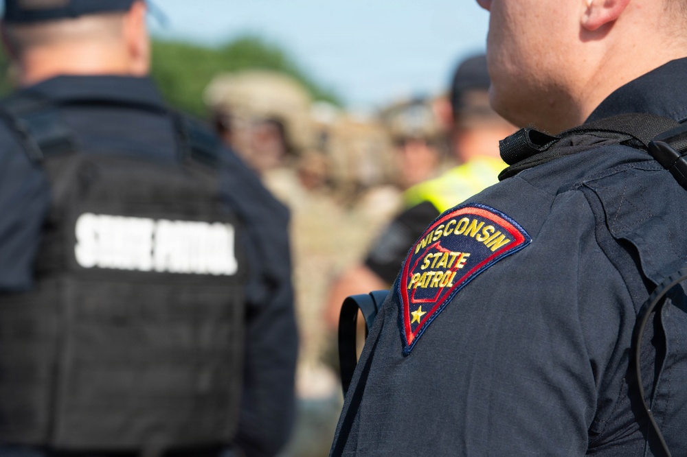 The image shows law enforcement personnel from the Wisconsin State Patrol gathered outdoors. The foreground features a close-up of an officer's shoulder patch reading 'Wisconsin State Patrol' in red, gold, and blue. In the background, multiple officers and individuals in various uniforms are visible, suggesting a multi-agency gathering or briefing.