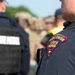 The image shows law enforcement personnel from the Wisconsin State Patrol gathered outdoors. The foreground features a close-up of an officer's shoulder patch reading 'Wisconsin State Patrol' in red, gold, and blue. In the background, multiple officers and individuals in various uniforms are visible, suggesting a multi-agency gathering or briefing.