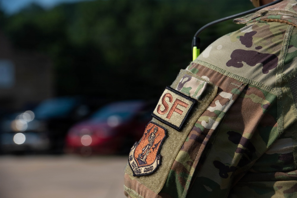 Close-up photograph of a military uniform sleeve showing unit patches on an OCP (Operational Camouflage Pattern) uniform. The patches identify the wearer as a Security Forces (SF) member of the Air National Guard. A radio antenna is visible over the shoulder, and blurred vehicles and trees appear in the background.