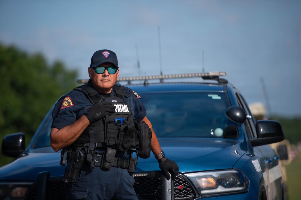 A Wisconsin State Patrol officer stands in front of a blue patrol vehicle in an outdoor setting. The officer is wearing full tactical gear including a vest marked 'PATROL' with badge number 2835, dark sunglasses, and a department cap. The image appears to be a professional or promotional photograph taken in good lighting conditions.