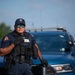 A Wisconsin State Patrol officer stands in front of a blue patrol vehicle in an outdoor setting. The officer is wearing full tactical gear including a vest marked 'PATROL' with badge number 2835, dark sunglasses, and a department cap. The image appears to be a professional or promotional photograph taken in good lighting conditions.