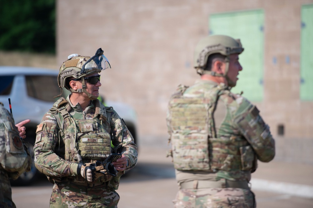 Two military personnel in full tactical gear are standing in an outdoor area, appearing to be engaged in a briefing or observation during a training exercise. Both are wearing OCP (Operational Camouflage Pattern) uniforms with plate carriers, tactical helmets, and other combat equipment. A concrete block building with green-marked panels is visible in the background, suggesting a training facility or range.