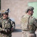 Two military personnel in full tactical gear are standing in an outdoor area, appearing to be engaged in a briefing or observation during a training exercise. Both are wearing OCP (Operational Camouflage Pattern) uniforms with plate carriers, tactical helmets, and other combat equipment. A concrete block building with green-marked panels is visible in the background, suggesting a training facility or range.