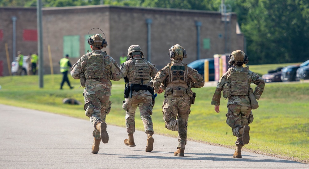 Four military personnel in full combat gear and camouflage uniforms are moving rapidly down a paved path toward a building. At least one individual has 'POLICE' marked on their back, indicating military police involvement. The scene appears to be a training exercise or active response situation on a military installation.