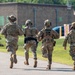 Four military personnel in full combat gear and camouflage uniforms are moving rapidly down a paved path toward a building. At least one individual has 'POLICE' marked on their back, indicating military police involvement. The scene appears to be a training exercise or active response situation on a military installation.