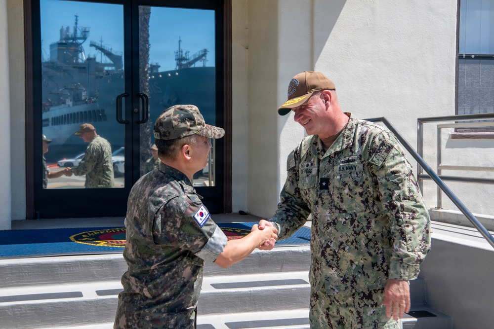 Two military personnel are shaking hands outside a building at what appears to be a naval installation. One individual wears a Republic of Korea (ROK) Army uniform with a South Korean flag patch, while the other wears a U.S. Navy uniform with a single star insignia indicating the rank of Rear Admiral (Lower Half). A naval vessel is visible in the background through the glass doors.