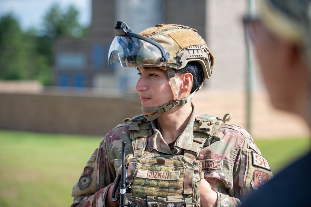 A U.S. Air Force Security Forces member named Guzman is pictured in full tactical gear, including a camouflage helmet with a mounted visor and a plate carrier vest. The service member appears to be engaged in a briefing or conversation with another individual partially visible in the foreground. The setting appears to be an outdoor area on a military installation.