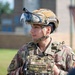 A U.S. Air Force Security Forces member named Guzman is pictured in full tactical gear, including a camouflage helmet with a mounted visor and a plate carrier vest. The service member appears to be engaged in a briefing or conversation with another individual partially visible in the foreground. The setting appears to be an outdoor area on a military installation.