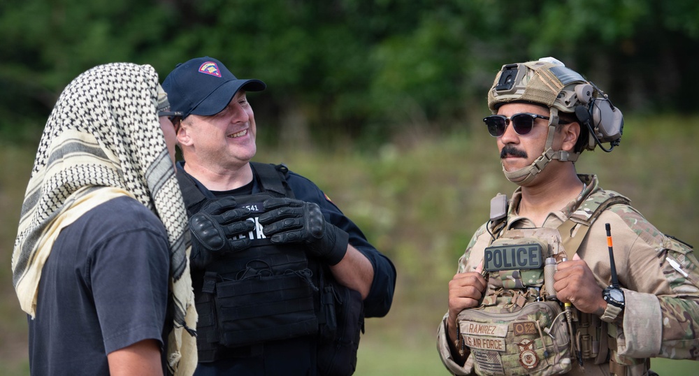 The image shows three individuals in what appears to be a military or law enforcement training exercise. One person wears a keffiyeh head covering, a second wears a black tactical vest with a baseball cap, and a third is in full U.S. Air Force military police gear including a tactical helmet and 'POLICE' patch. The individuals appear relaxed and conversational, suggesting a break or debrief during a training scenario.