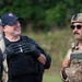 The image shows three individuals in what appears to be a military or law enforcement training exercise. One person wears a keffiyeh head covering, a second wears a black tactical vest with a baseball cap, and a third is in full U.S. Air Force military police gear including a tactical helmet and 'POLICE' patch. The individuals appear relaxed and conversational, suggesting a break or debrief during a training scenario.