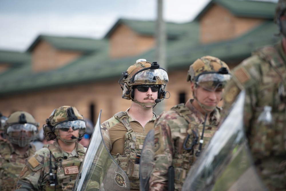 The image shows a group of military personnel in full tactical gear, including helmets with mounted equipment, communication headsets, sunglasses, and camouflage uniforms. They appear to be holding transparent riot shields, suggesting participation in a crowd control or civil disturbance training exercise. The setting appears to be a military installation or training facility with brick buildings visible in the background.