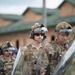The image shows a group of military personnel in full tactical gear, including helmets with mounted equipment, communication headsets, sunglasses, and camouflage uniforms. They appear to be holding transparent riot shields, suggesting participation in a crowd control or civil disturbance training exercise. The setting appears to be a military installation or training facility with brick buildings visible in the background.