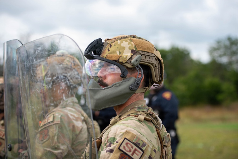 A military Security Forces (SF) personnel is shown in profile view wearing full tactical gear including a multicam helmet with face shield and chin guard. The individual has face paint applied and is holding or standing behind a clear riot/ballistic shield, suggesting participation in a training exercise. Additional personnel in similar gear and what appears to be a role-player in a blue shirt are visible in the background.