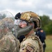 A military Security Forces (SF) personnel is shown in profile view wearing full tactical gear including a multicam helmet with face shield and chin guard. The individual has face paint applied and is holding or standing behind a clear riot/ballistic shield, suggesting participation in a training exercise. Additional personnel in similar gear and what appears to be a role-player in a blue shirt are visible in the background.