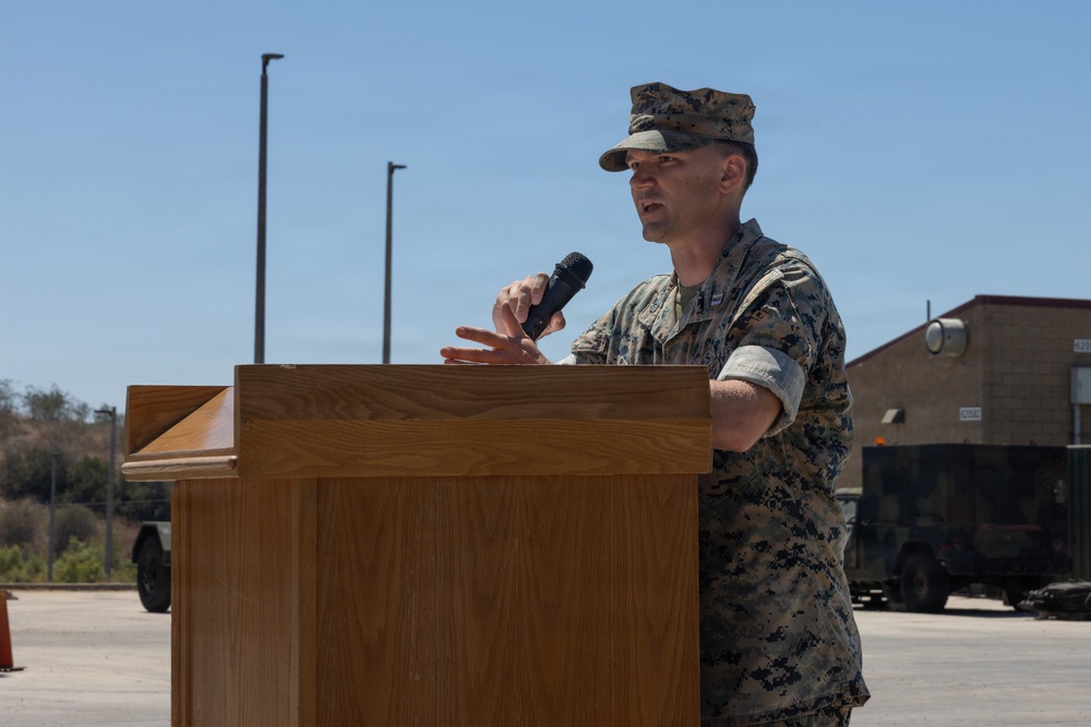 A military service member in camouflage uniform is delivering a speech at a wooden podium while holding a handheld microphone. The setting appears to be an outdoor ceremony or briefing on what looks like a military installation. The background shows military vehicles, utility poles, and a building consistent with a base environment.