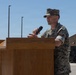 A military service member in camouflage uniform is delivering a speech at a wooden podium while holding a handheld microphone. The setting appears to be an outdoor ceremony or briefing on what looks like a military installation. The background shows military vehicles, utility poles, and a building consistent with a base environment.