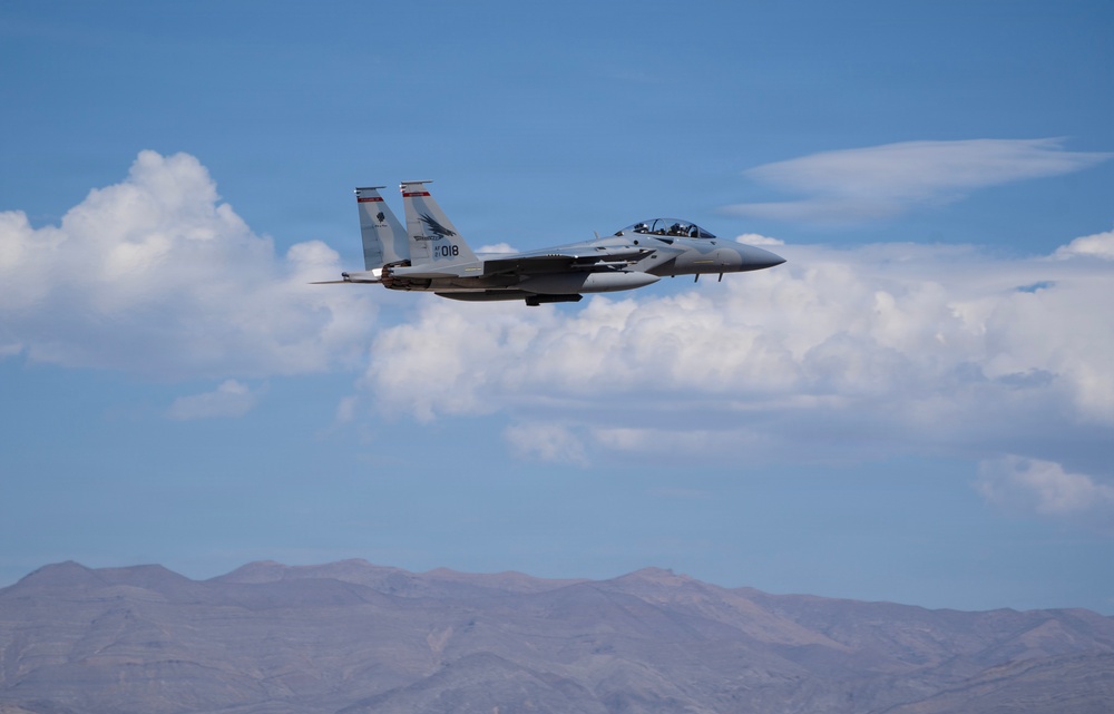 A U.S. Air Force F-15 Eagle fighter jet, marked with tail number '018', is captured in level flight against a partly cloudy blue sky. The aircraft appears to be carrying external stores/weapons and is flying over a mountainous desert terrain. The image is sharp and well-composed, suggesting it was taken by a professional or experienced photographer.