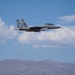 A U.S. Air Force F-15 Eagle fighter jet, marked with tail number '018', is captured in level flight against a partly cloudy blue sky. The aircraft appears to be carrying external stores/weapons and is flying over a mountainous desert terrain. The image is sharp and well-composed, suggesting it was taken by a professional or experienced photographer.
