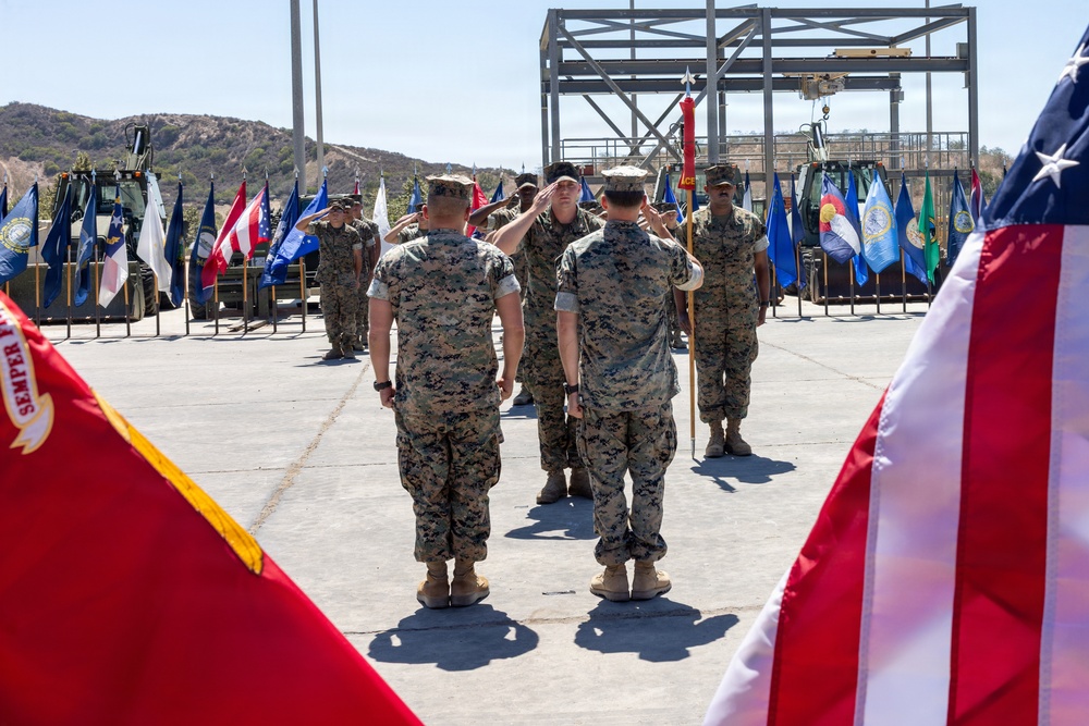 A formal U.S. Marine Corps military ceremony is taking place on an outdoor paved area, likely a parade deck at a military installation. Multiple Marines in MARPAT camouflage uniforms are rendering salutes, with numerous state and military flags displayed in the background. The American flag and a Marine Corps flag with 'Semper Fi' visible are prominently framing the foreground.