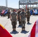 A formal U.S. Marine Corps military ceremony is taking place on an outdoor paved area, likely a parade deck at a military installation. Multiple Marines in MARPAT camouflage uniforms are rendering salutes, with numerous state and military flags displayed in the background. The American flag and a Marine Corps flag with 'Semper Fi' visible are prominently framing the foreground.