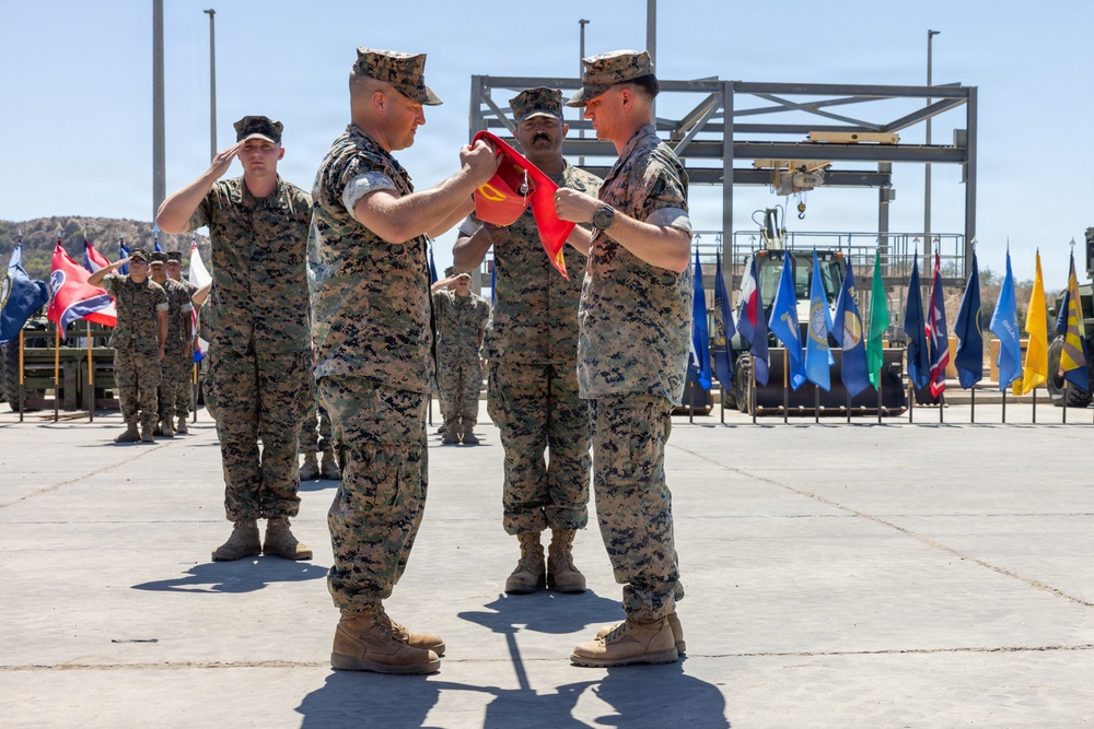 A military ceremony is taking place outdoors on a paved surface, likely a change of command or unit activation/deactivation ceremony. Two Marines in the foreground are handling a red guidon or unit flag, while other personnel stand at attention or salute in the background. Multiple state and unit flags are displayed on flag poles in the background.