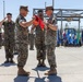 A military ceremony is taking place outdoors on a paved surface, likely a change of command or unit activation/deactivation ceremony. Two Marines in the foreground are handling a red guidon or unit flag, while other personnel stand at attention or salute in the background. Multiple state and unit flags are displayed on flag poles in the background.