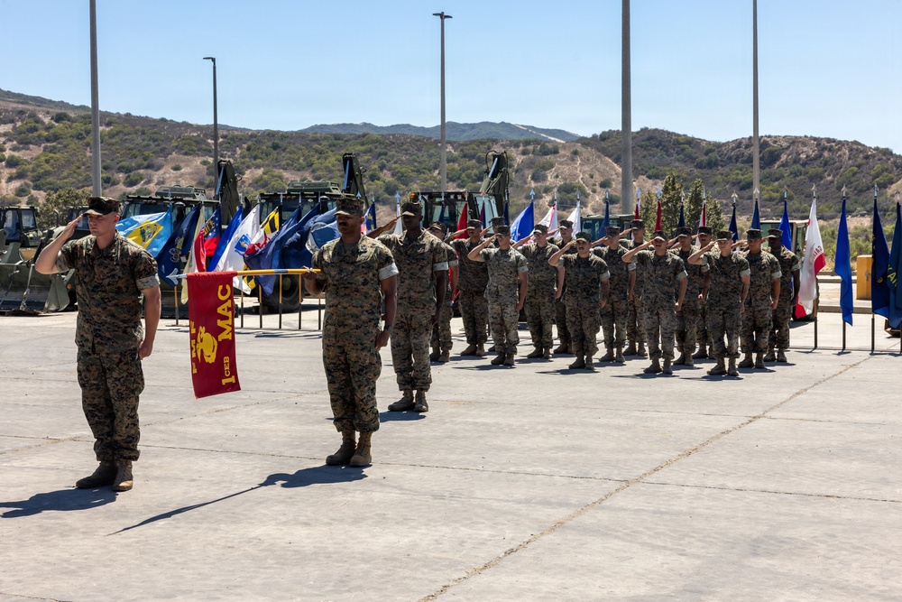 A formal U.S. Marine Corps military ceremony is taking place outdoors on a paved parade ground. Personnel in MARPAT camouflage uniforms are standing in formation and rendering salutes, with a guidon bearer holding a red unit flag marked '1 CEB MAC'. Multiple unit and organizational flags are displayed in the background alongside military construction equipment and hilly terrain.