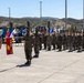 A formal U.S. Marine Corps military ceremony is taking place outdoors on a paved parade ground. Personnel in MARPAT camouflage uniforms are standing in formation and rendering salutes, with a guidon bearer holding a red unit flag marked '1 CEB MAC'. Multiple unit and organizational flags are displayed in the background alongside military construction equipment and hilly terrain.