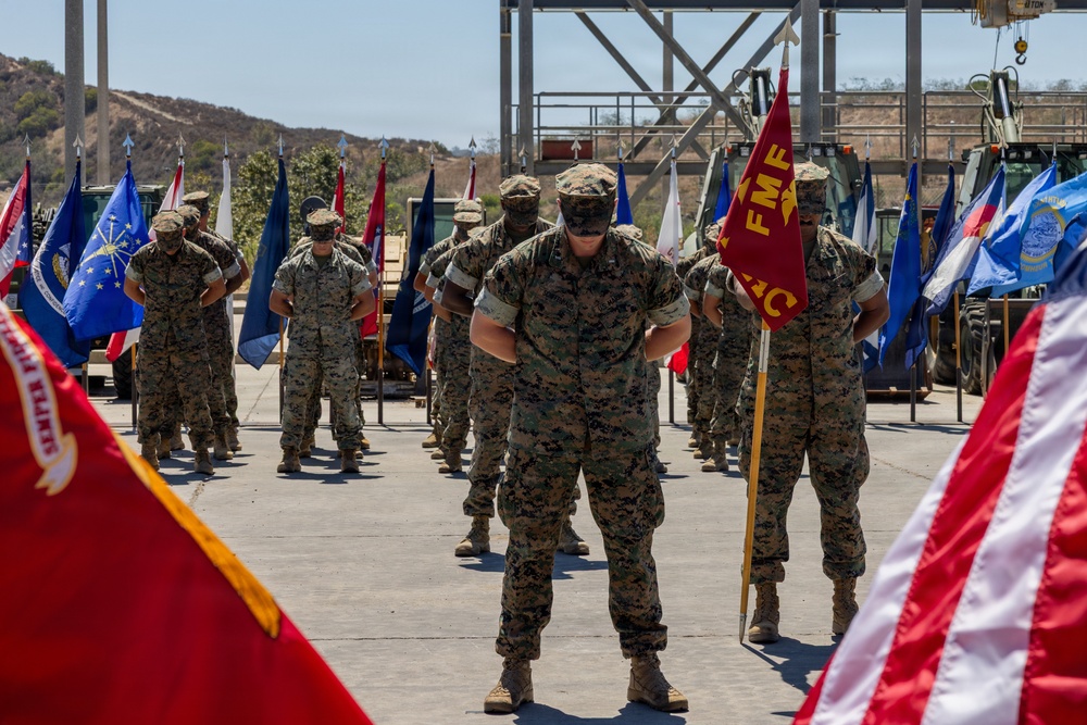 A formal U.S. Marine Corps military ceremony is taking place outdoors on a paved surface, likely at a military installation. Multiple Marines in woodland MARPAT camouflage uniforms stand at parade rest in organized rows, flanked by numerous unit and state flags. A red FMF (Fleet Marine Force) guidon is prominently visible among the color guard.
