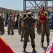 A formal U.S. Marine Corps military ceremony is taking place outdoors on a paved surface, likely at a military installation. Multiple Marines in woodland MARPAT camouflage uniforms stand at parade rest in organized rows, flanked by numerous unit and state flags. A red FMF (Fleet Marine Force) guidon is prominently visible among the color guard.