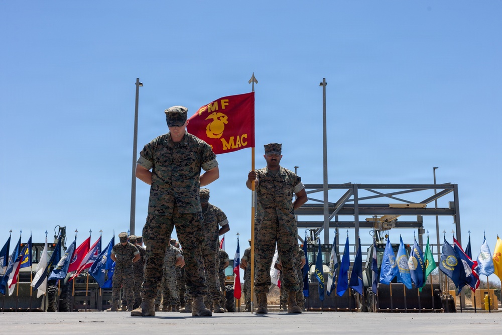 A U.S. Marine Corps military ceremony is taking place outdoors, featuring Marines in woodland MARPAT camouflage uniforms standing in formation. A guidon bearer holds a red FMF MAC (Fleet Marine Force, Marine Air-Ground Task Force Command) flag prominently in the foreground. Multiple state and unit flags are displayed in the background alongside construction equipment and flag poles.