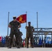 A U.S. Marine Corps military ceremony is taking place outdoors, featuring Marines in woodland MARPAT camouflage uniforms standing in formation. A guidon bearer holds a red FMF MAC (Fleet Marine Force, Marine Air-Ground Task Force Command) flag prominently in the foreground. Multiple state and unit flags are displayed in the background alongside construction equipment and flag poles.