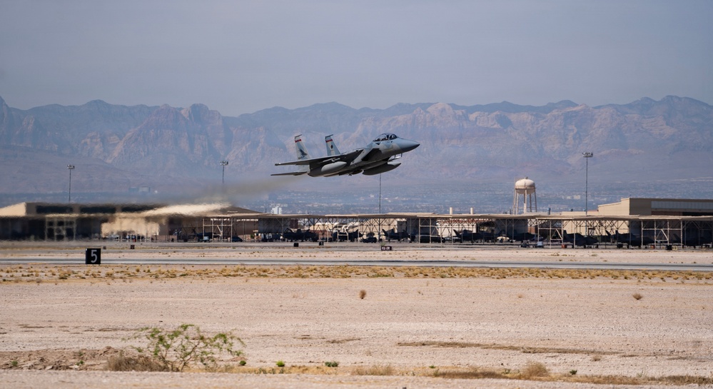 A military fighter jet, identifiable as an F-15 Eagle, is captured during takeoff from a desert airbase. The aircraft is in a steep climb with afterburner exhaust visible, set against a backdrop of arid terrain and distant mountain ranges. Ground infrastructure including hangars, a water tower, and runway markings are visible in the background.