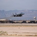A military fighter jet, identifiable as an F-15 Eagle, is captured during takeoff from a desert airbase. The aircraft is in a steep climb with afterburner exhaust visible, set against a backdrop of arid terrain and distant mountain ranges. Ground infrastructure including hangars, a water tower, and runway markings are visible in the background.